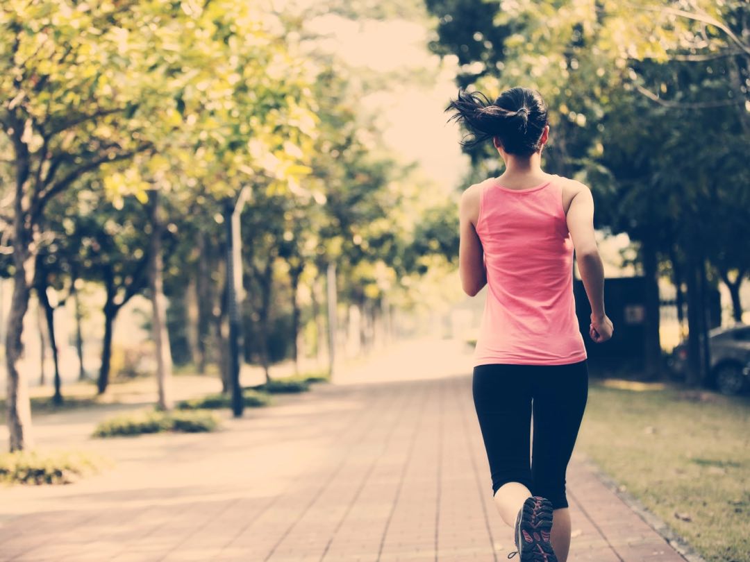 A woman with earbuds in, jogging along a tree-lined path